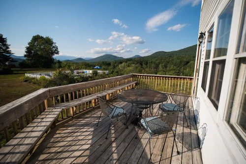 A wooden deck with a round table and chairs overlooks a scenic view of mountains and greenery under a blue sky.