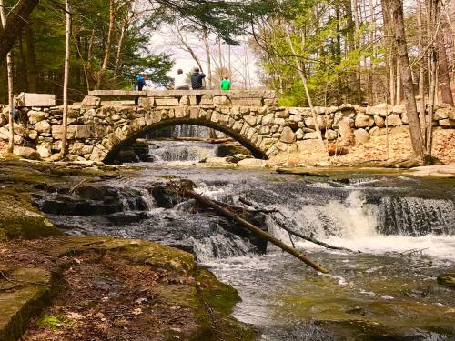 A stone bridge arches over a flowing stream, surrounded by trees and rocky terrain, with people walking on the bridge.