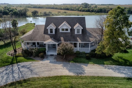 Aerial view of a large, charming house with a wraparound porch, surrounded by greenery and near a calm lake.