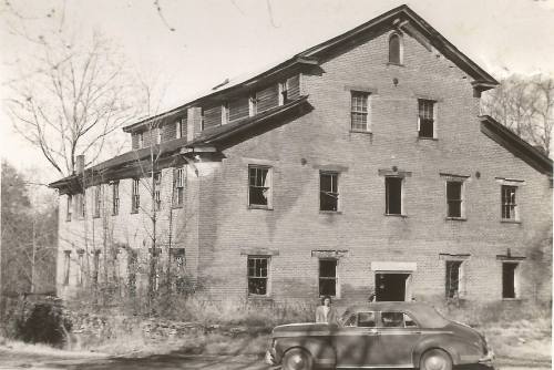 Black and white photo of an old brick building with broken windows and a vintage car parked nearby.