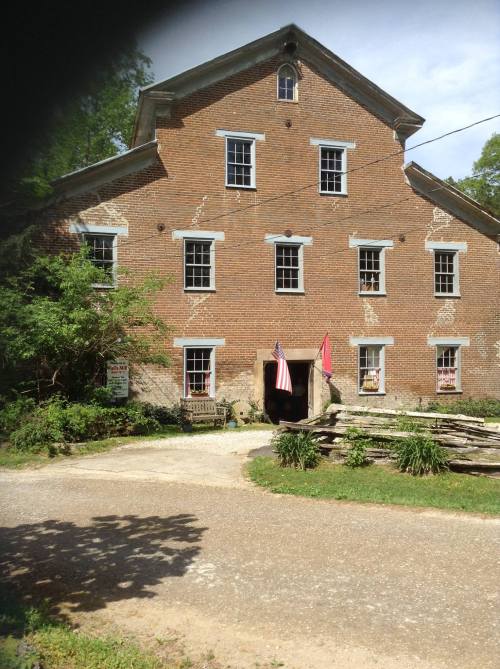 Historic brick building with multiple windows, flags hanging, surrounded by greenery and a gravel path.