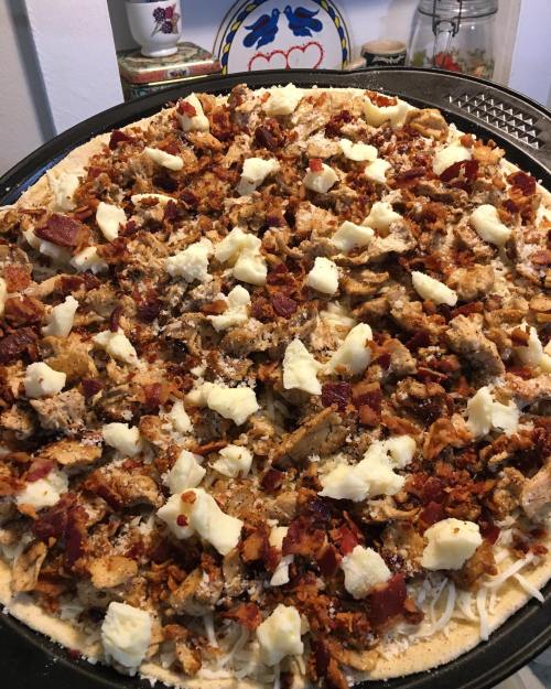 A close-up of a pizza topped with shredded chicken, bacon bits, and chunks of cheese on a golden crust.