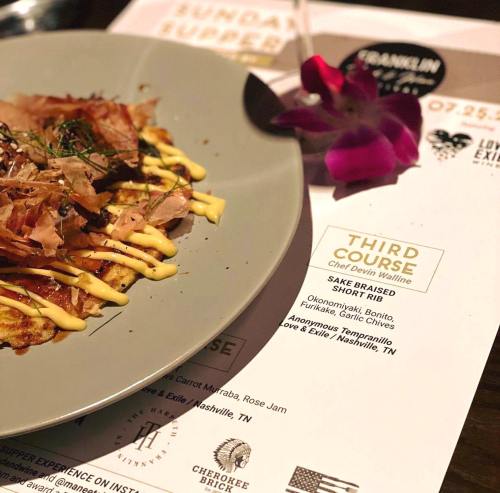 A plate of food featuring a savory dish topped with bonito flakes and drizzled with sauce, accompanied by an orchid.