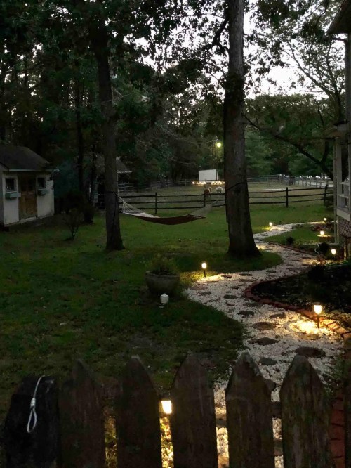 A serene backyard at dusk, featuring a hammock between trees, a winding path with lights, and a wooden fence.