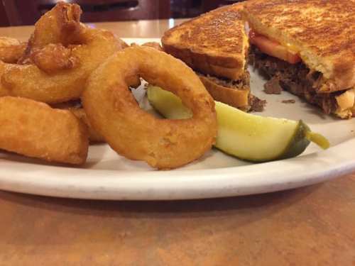 A plate featuring a grilled sandwich with tomato and pickles, alongside crispy onion rings.