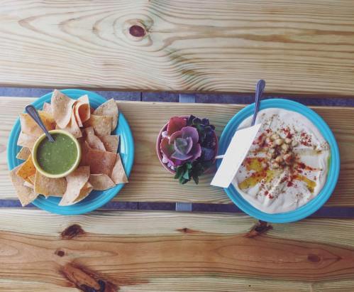 A wooden table with two plates: one with tortilla chips and green salsa, the other with creamy dip and garnishes.