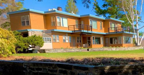 A modern two-story house with a brown exterior, blue trim, and balconies, surrounded by greenery and a clear blue sky.