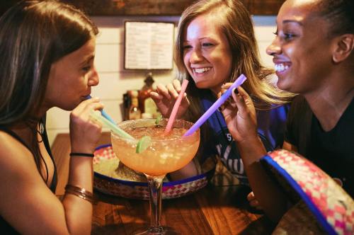 Three women share a large cocktail, laughing and sipping through colorful straws at a lively restaurant.