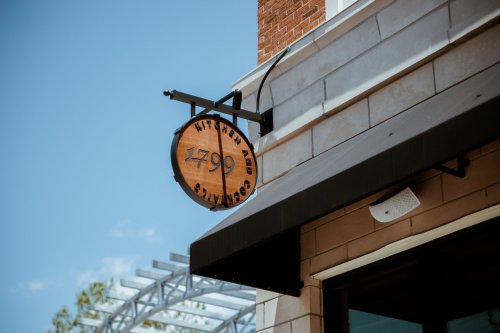 Sign for "1799 Kitchen and Cocktails" hanging from a building, with a clear blue sky in the background.