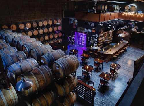 A spacious brewery interior with wooden barrels, a bar, tables, and ambient lighting.