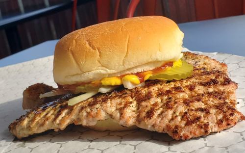 A grilled fish fillet topped with a hamburger bun, garnished with ketchup, mustard, and pickles on a paper tray.