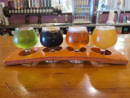 A flight of four small beer glasses on a wooden paddle, featuring green, black, amber, and golden brews.