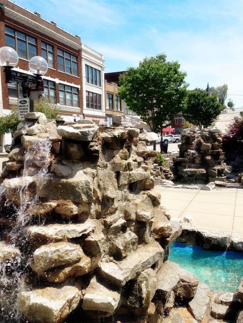A stone fountain with flowing water, surrounded by greenery and buildings under a clear blue sky.
