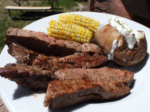 A plate of grilled steak slices, corn on the cob, and a baked potato topped with sour cream and chives.