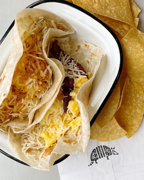 Three breakfast tacos filled with eggs, cheese, and potatoes, served with crispy tortilla chips on the side.