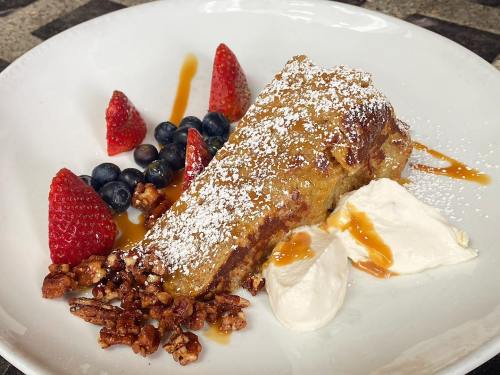 A slice of French toast topped with powdered sugar, served with strawberries, blueberries, caramel sauce, and whipped cream.