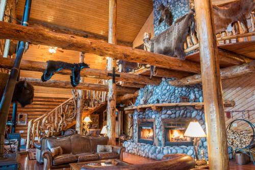 Cozy log cabin interior with stone fireplace, wooden beams, and rustic decor, featuring animal hides and warm lighting.