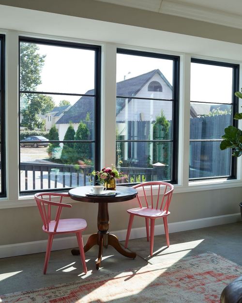 A cozy dining nook with a round table and two pink chairs, surrounded by large windows and a view of greenery outside.