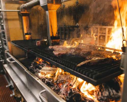 A grill with steaks cooking over an open flame, surrounded by glowing embers in a restaurant kitchen.