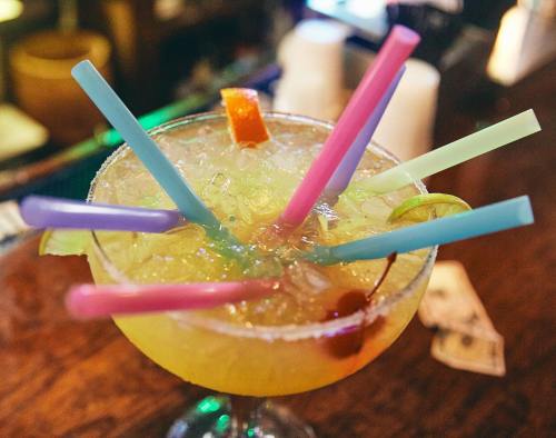 A colorful cocktail with multiple straws, garnished with a cherry and orange slice, served in a glass on a wooden bar.