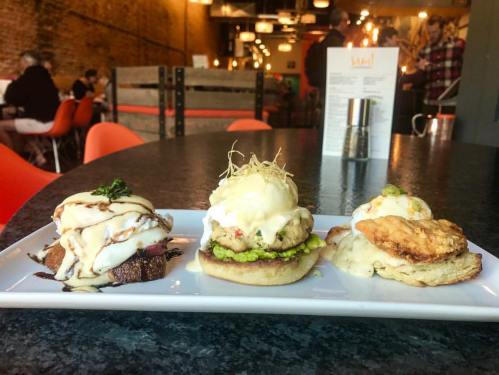 A plate with three gourmet brunch dishes, featuring eggs, avocado, and sauces, set on a dark table in a cozy restaurant.