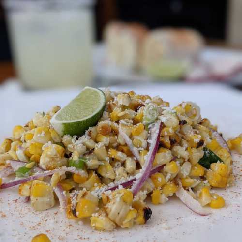 A vibrant corn salad with lime, red onion, and spices, served on a white plate with a drink in the background.