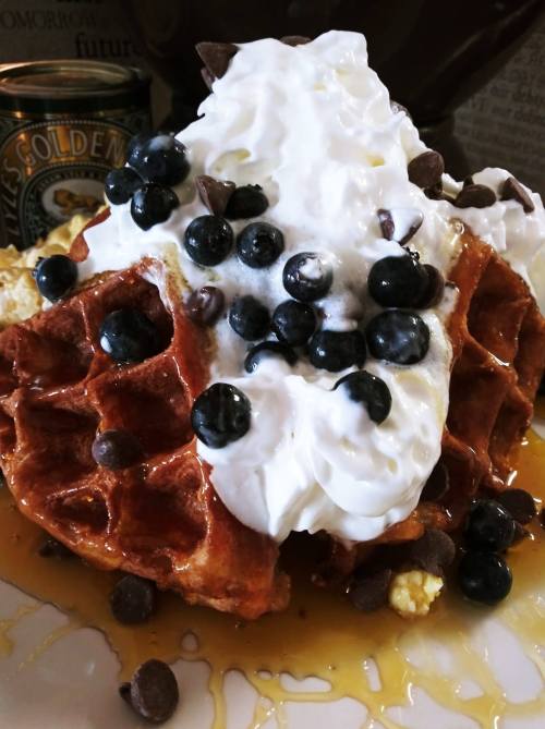 A close-up of a waffle topped with whipped cream, blueberries, chocolate chips, and drizzled with syrup.