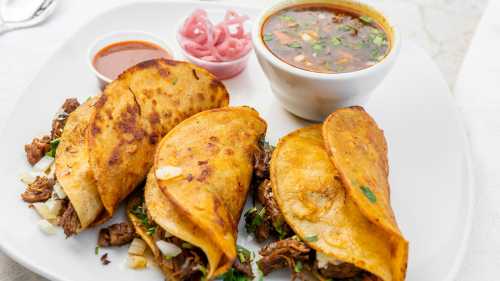 Three tacos filled with meat and onions, served with a bowl of broth, pickled onions, and a side of sauce.