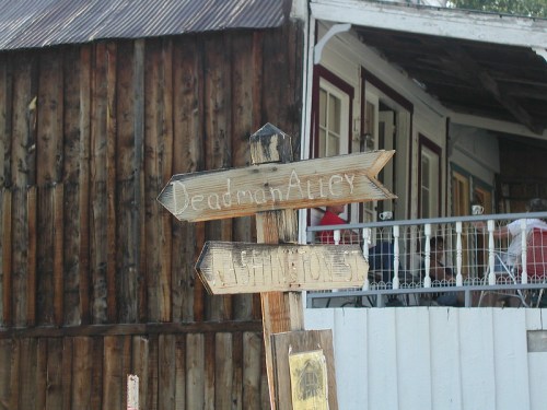 Wooden signpost with arrows pointing to "Deadman Alley" and "Cashin Stones," in front of a rustic building.