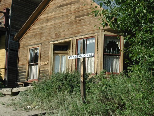 A weathered wooden house with lace curtains and a street sign reading "Washington St" in a grassy area.