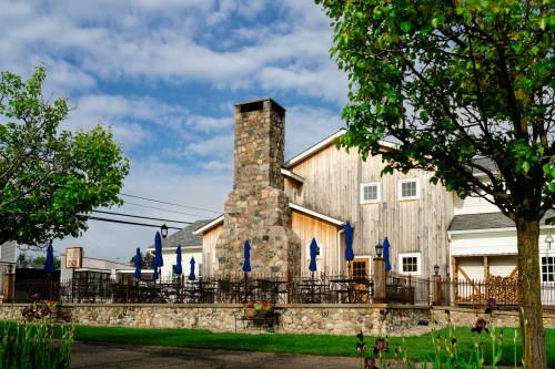 A rustic building with a stone chimney, surrounded by trees and outdoor seating under blue umbrellas.