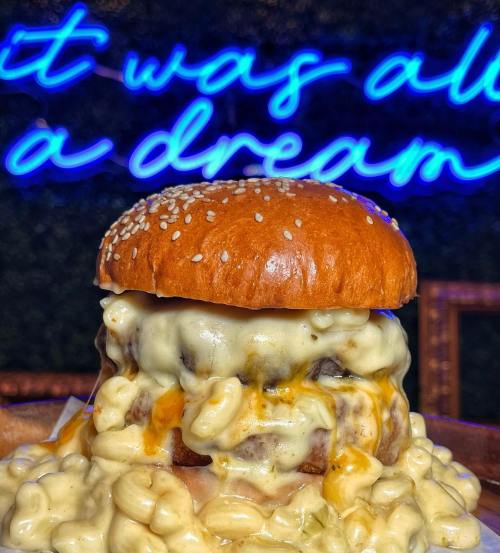 A close-up of a cheesy burger topped with macaroni and cheese, set against a neon sign that reads "It was all a dream."