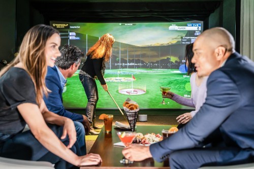 A group of friends enjoying a virtual golf game on a large screen, with drinks and snacks on the table.