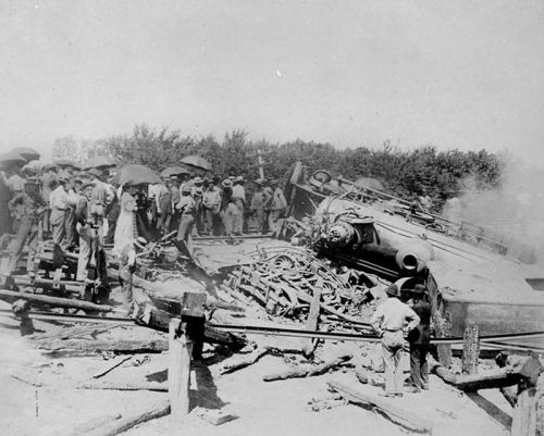 A crowd gathers around a derailed train, with debris scattered and smoke rising in a historical black-and-white photo.