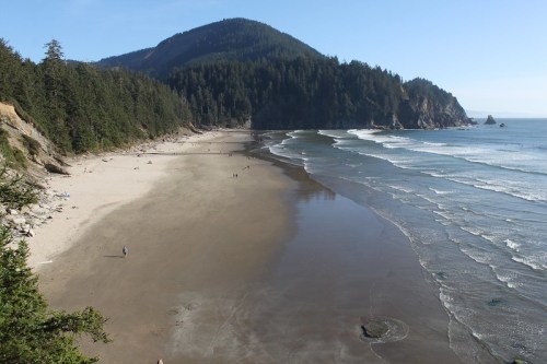 A scenic beach with gentle waves, surrounded by lush green hills and a clear blue sky. People stroll along the shore.