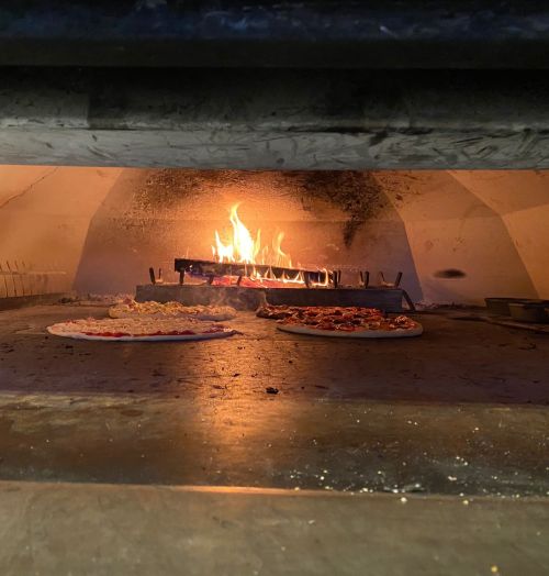 A wood-fired oven with pizzas baking inside, flames visible in the background.