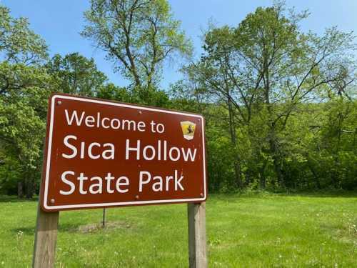 A brown sign reading "Welcome to Sica Hollow State Park" in a green, wooded area on a sunny day.