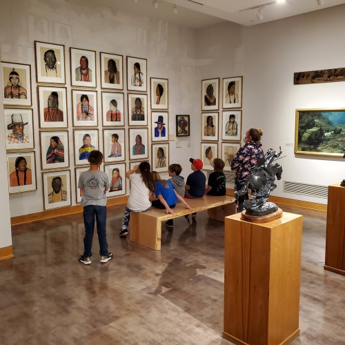 Children and adults observe framed portraits in an art gallery, seated on a wooden bench.