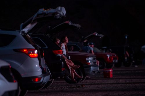 A woman and child sit on the tailgate of a car, enjoying a night view in a parking lot filled with vehicles.