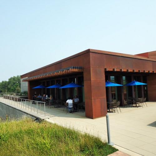A modern building with a copper facade and outdoor seating under blue umbrellas on a sunny day.
