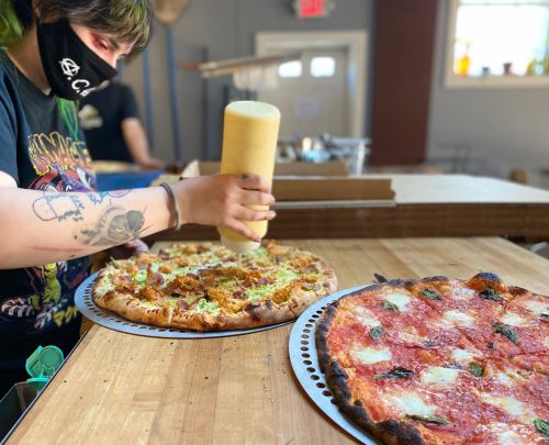 A person adds sauce to a pizza on a wooden table, with another pizza nearby, in a casual kitchen setting.