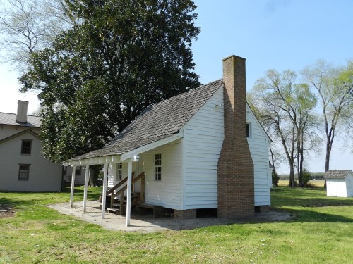 A small white house with a chimney, surrounded by grass and trees, featuring a porch and a simple design.