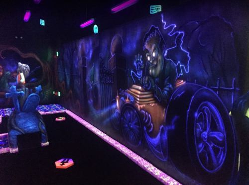 A vibrant, neon-lit mini-golf course featuring spooky murals and glowing obstacles in a dark environment.