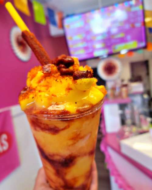 A colorful dessert cup filled with mango, chili powder, and a cinnamon stick, set against a vibrant shop background.