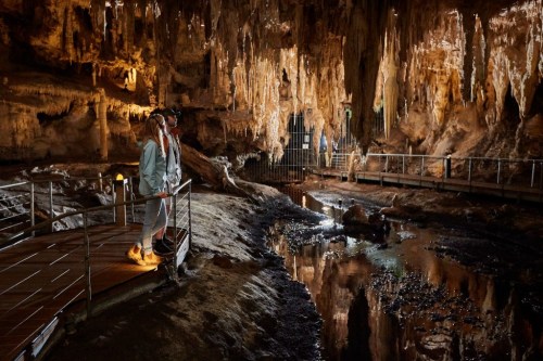 A person stands on a wooden walkway in a cave, surrounded by stalactites and a reflective pool of water.