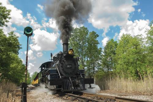A vintage steam train emits black smoke as it travels along a track surrounded by lush greenery and a blue sky.