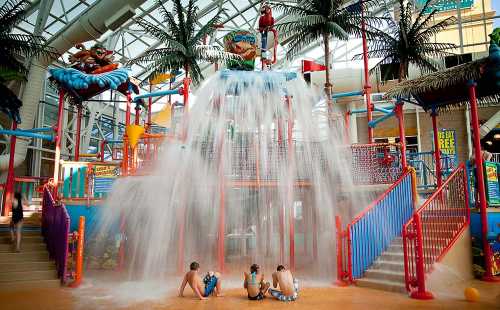 Children playing at a water park with a large water feature and colorful structures under a glass roof.