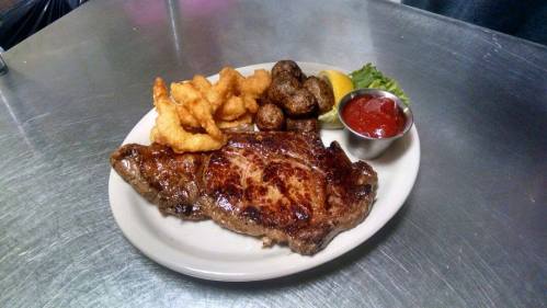 A plate of grilled steak with shrimp, roasted potatoes, a lemon wedge, lettuce, and a side of ketchup.