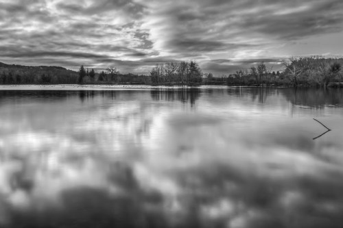 A serene black and white landscape of a calm lake reflecting cloudy skies and surrounding trees.