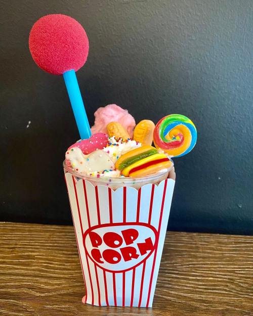A colorful dessert in a popcorn box, topped with whipped cream, candy, a lollipop, and a pink cotton candy ball.